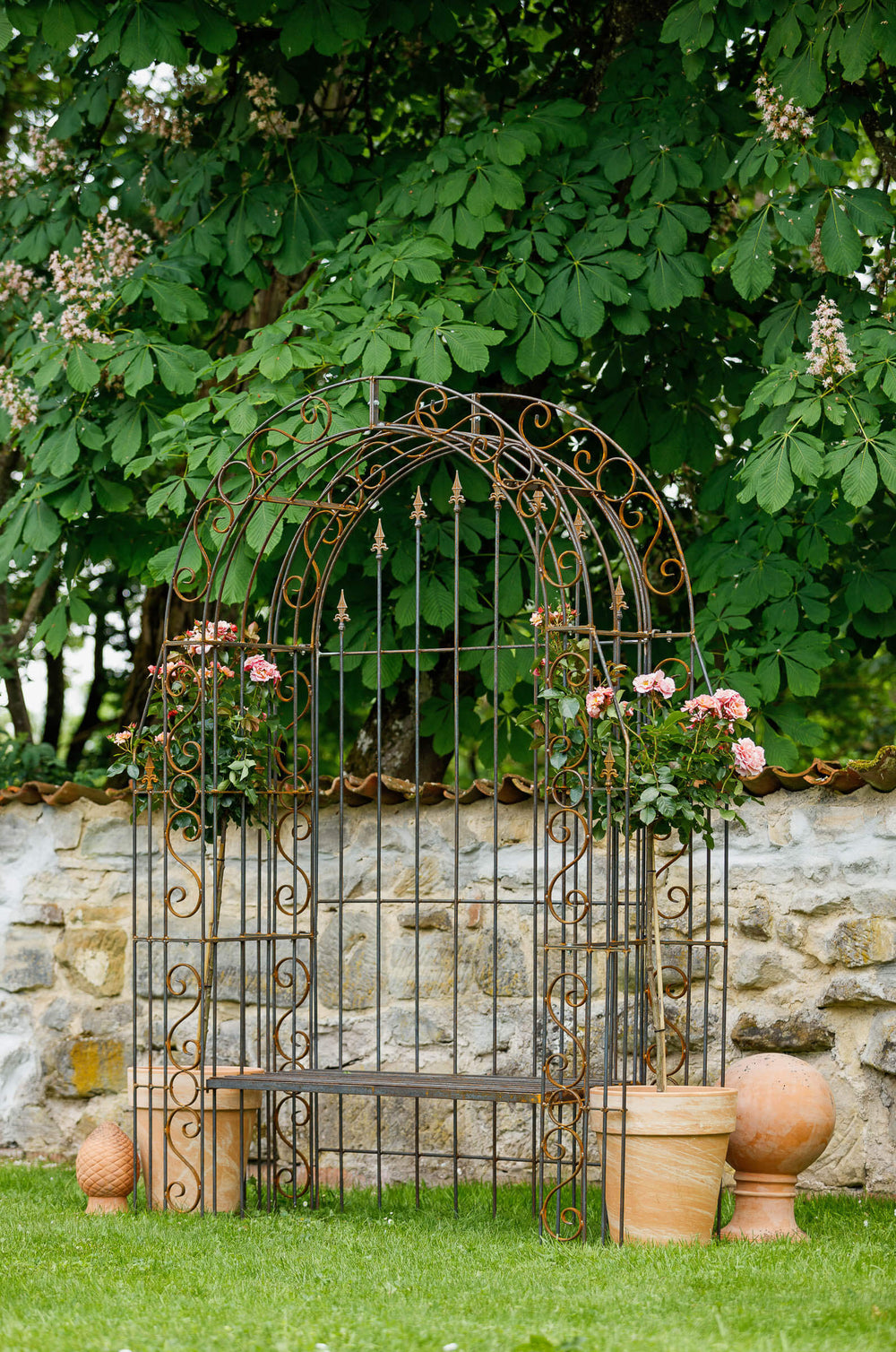 Rosenbogen Calabria mit Bank und Rückwand in Rostoptik Gesamtansicht mit Rosen und Dekoration vor Mauer und grünen Bäumen Rosenbogen Calabria mit Bank und Rückwand in Rostoptik Gesamtansicht mit Rosen und Dekoration vor Mauer und grünen Bäumen
